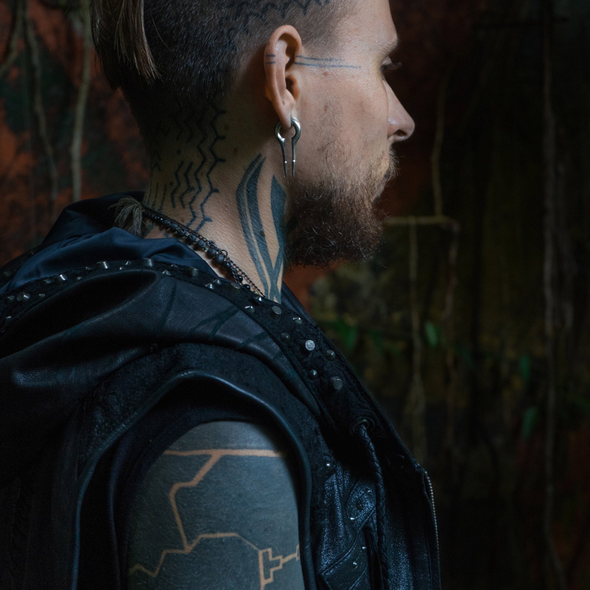 A person with a side-profile view showcases an elaborate undercut hairstyle with shaved patterns and tattoos on their neck and arm. They wear a leather vest adorned with metal studs and accessorize with multiple CALYPSO Minimalist Keyhole Ear Weights in 925 Silver from Kaya Movement, sized at 2 gauge. The background is dark with hints of greenery.
